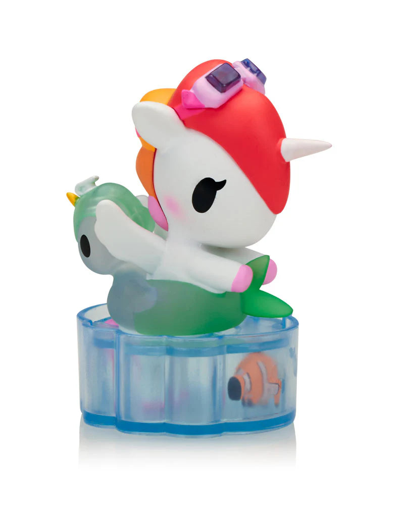 Fun In The Sun Unicorno Blind Box: White unicorn figurine with red-orange hair, pink goggles, on green float. Blue base holds a clownfish.