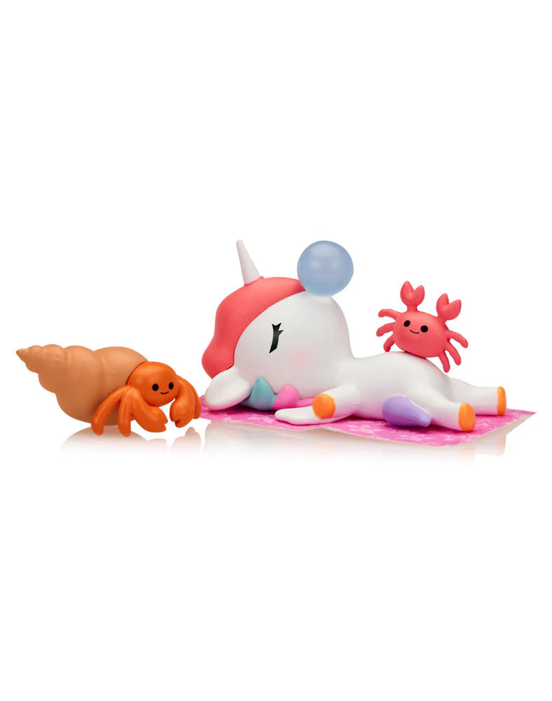 Fun In The Sun Unicorno Blind Box: Glossy unicorn-narwhal toy with pastel hooves, pink mat, blowing bubblegum, red crab on back, orange hermit-crab alongside. Collectible vinyl figurine.