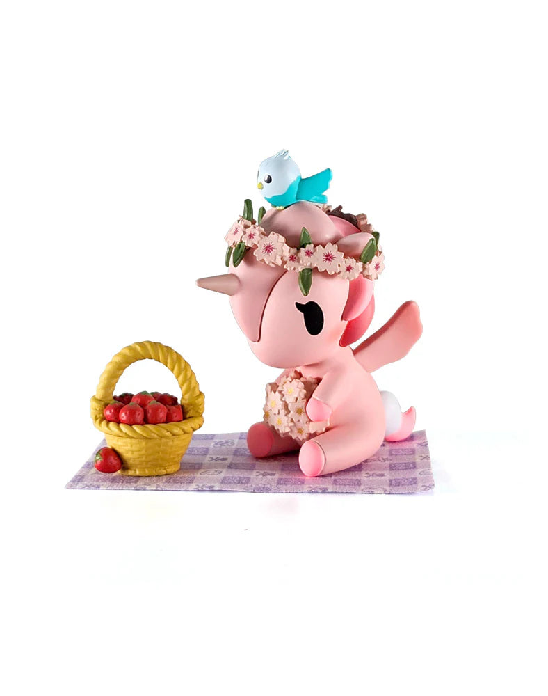 Pink winged unicorn from Picnic Party Unicorno Blind Box, adorned with cherry blossoms and a blue bird, sits beside a basket of strawberries.