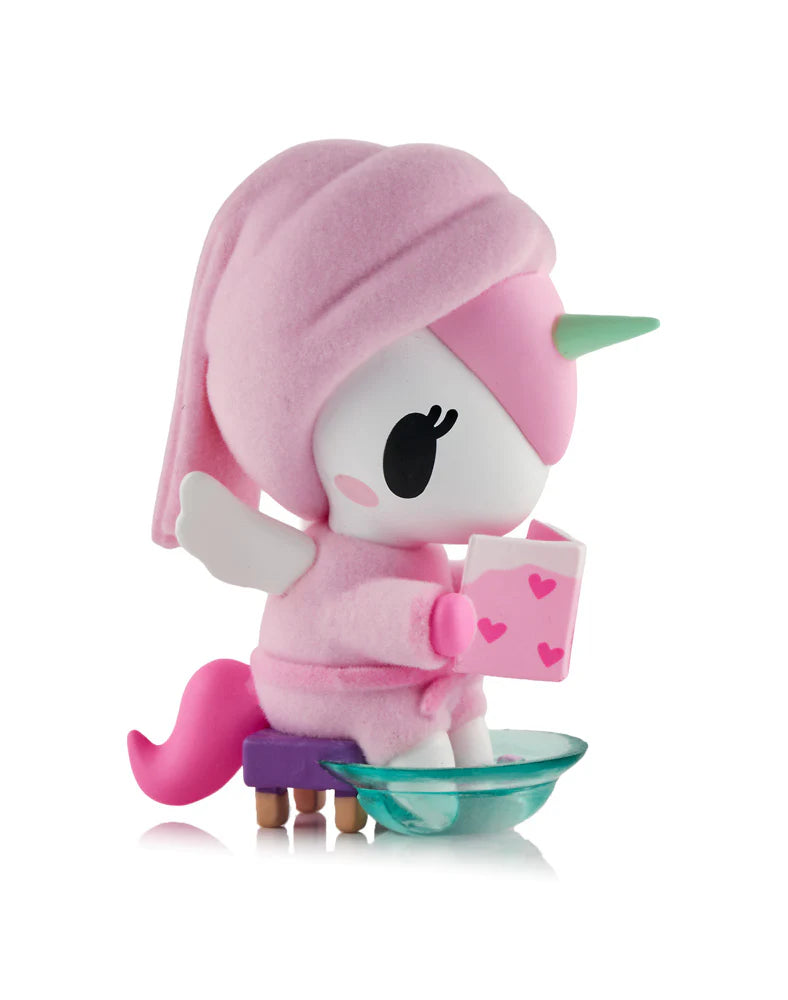 Spa Day Unicorno Blind Box figurine: a unicorn in a pink robe with a towel turban, seated on a stool, reading a heart-covered book.