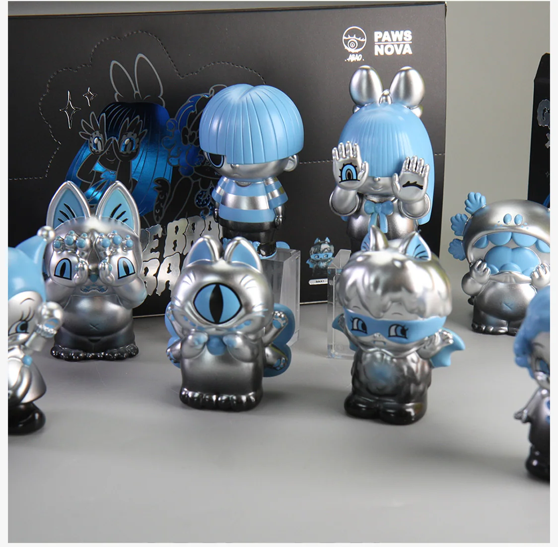 GRAPE BRAIN x ABAO BLINDBOX figures in blue and silver, featuring whimsical, cartoonish designs with oversized heads, in front of PAWS NOVA packaging.