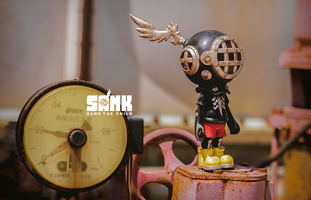 Miniature toy figurine of a person with a helmet and wings by SANK TOYS, placed on a rusty metal surface.