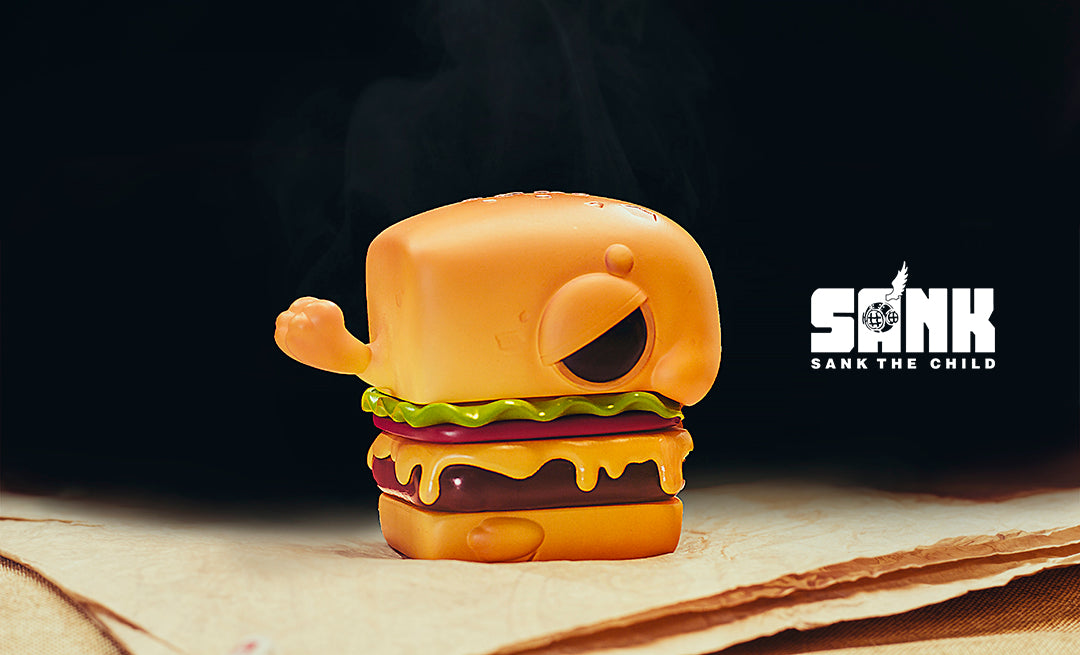 Hamburger Seal by Sank Toys, a toy burger with a yellow face on a table, made of resin, limited edition.