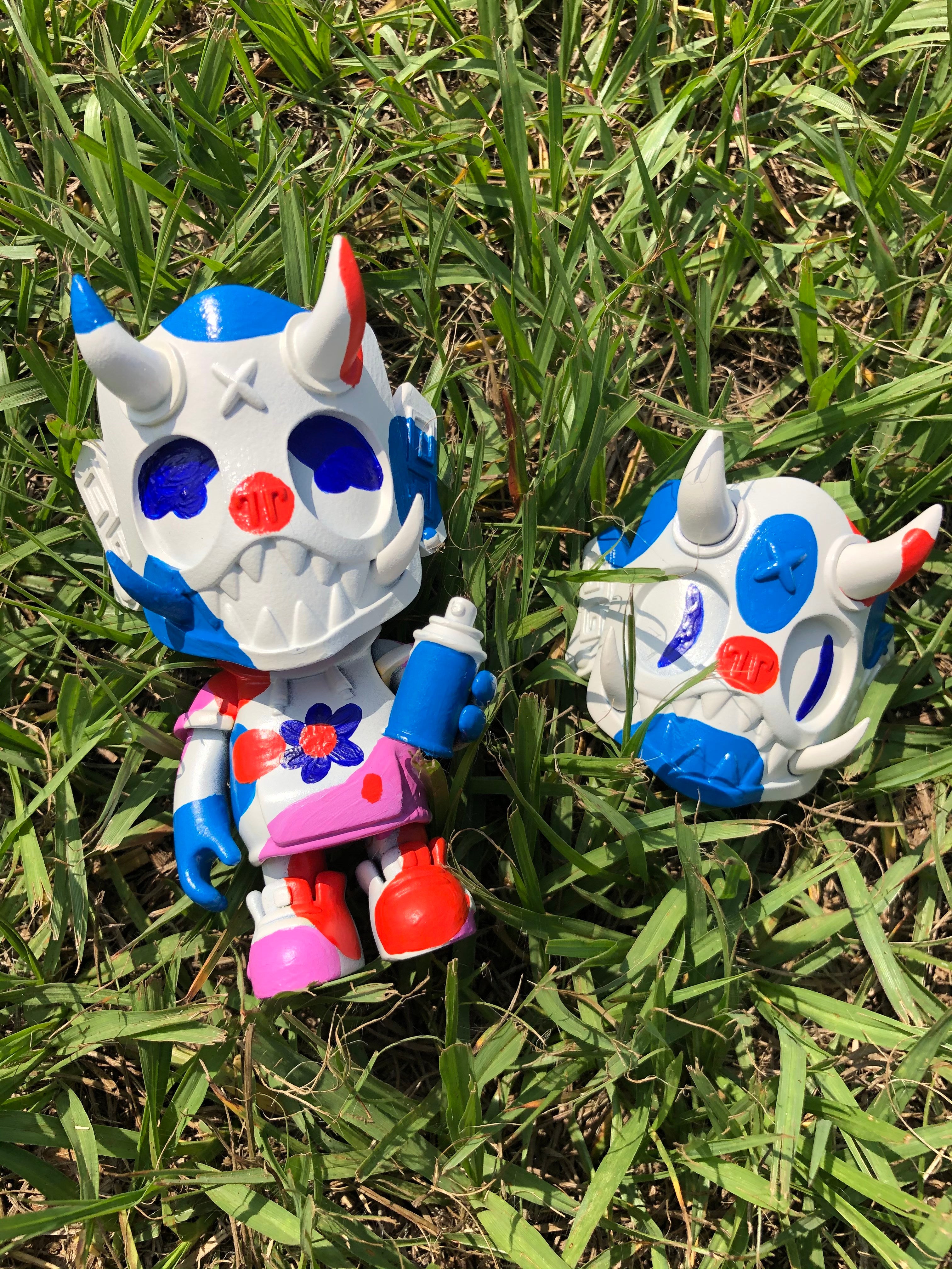 Two toy figures in the grass, one with a red circle and letter carved in it, part of Goblin Squad - Color flight by Jujmo.