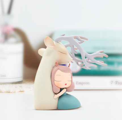 Figurine of a girl hugging a deer and cartoon character hugging a white animal, with a blurry image of a person's head and belly, and a cartoon animal with antlers.