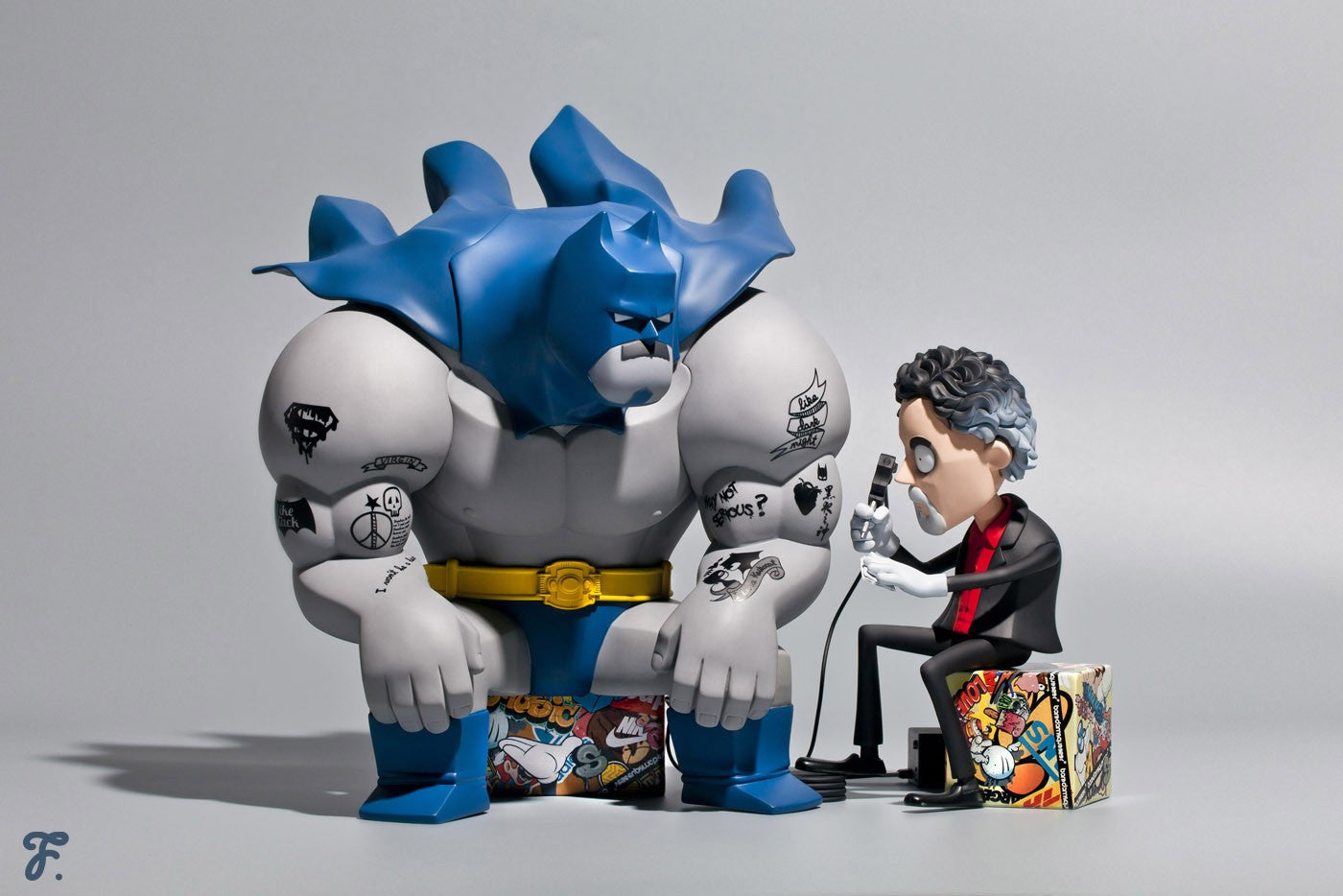 Toy figurine of superhero sitting on box with microphone, gorilla statue, cartoon character with power cord, and logo close-up.
